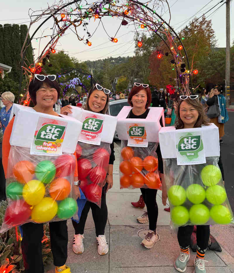 Four smiling women stand outdoors in creative tic tac costume outfits made from clear bags filled with balloons. They wear matching heart-shaped glasses and pose under a lit archway adorned with Halloween decorations.