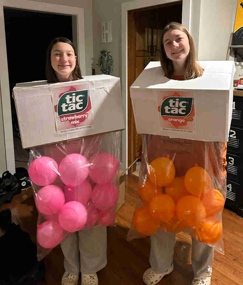Two people wear creative tic tac costumes—one labeled "strawberry" with pink balloons, the other "orange" with orange balloons—smiling and posing indoors in their fun homemade outfits.