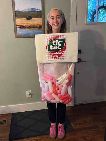 A girl smiles while wearing a creative tic tac costume made from a box and pink and white balloons, standing indoors on a wooden floor.