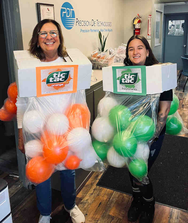 Two smiling women stand indoors in creative tic tac costumes made from boxes and plastic bags filled with orange, white, and green balloons, perfectly mimicking Tic Tac candy containers.