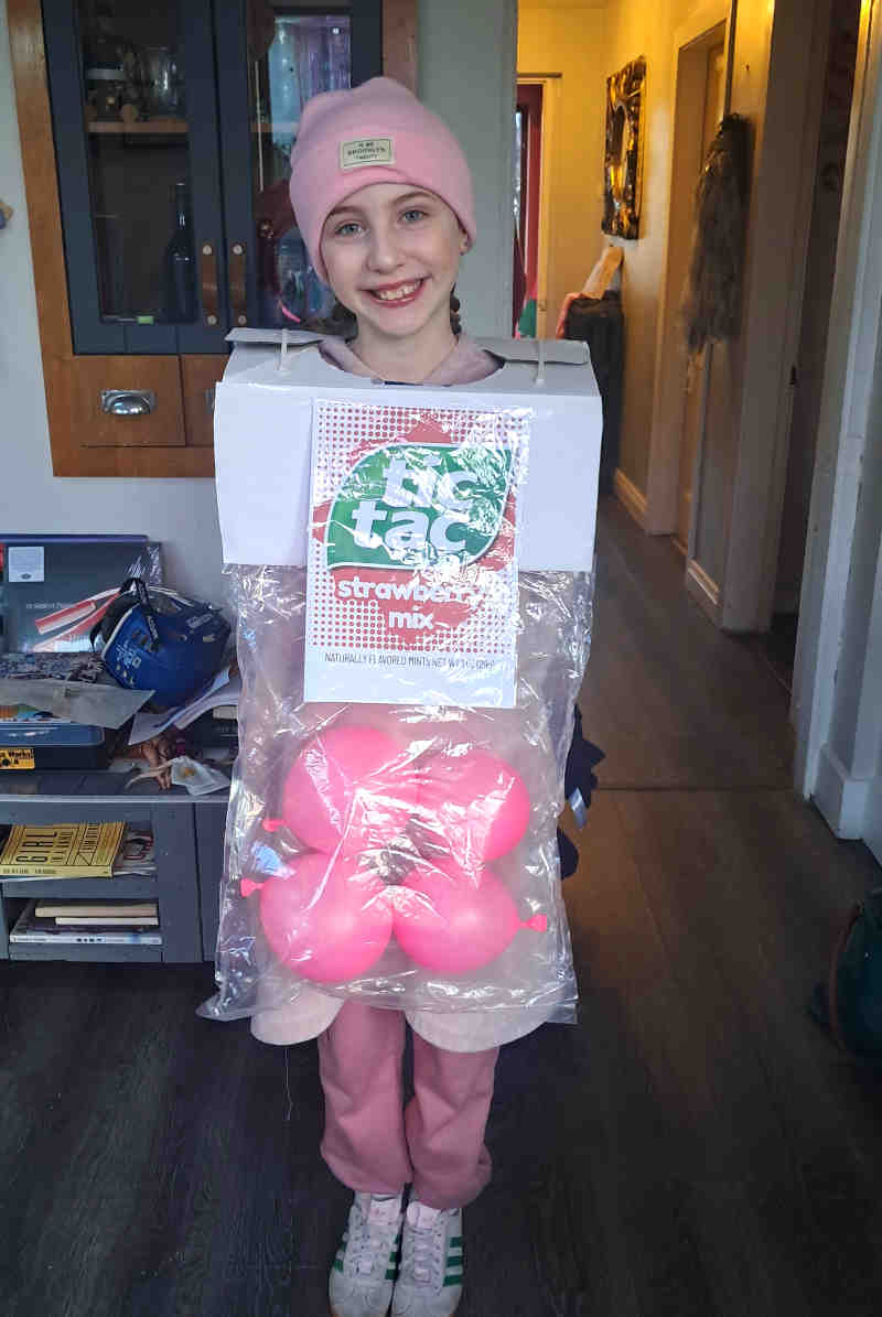 A smiling girl in a pink hat and pants is dressed in a homemade tic tac costume, styled as Strawberry Mix with pink balloons inside a box, standing in a warmly lit room.