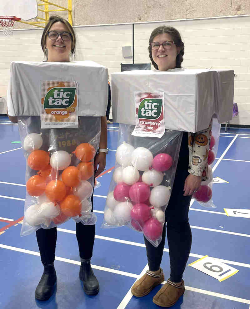 Two people smile while wearing creative Tic Tac costume boxes—one as orange, the other as strawberry-mix. The clever costumes use plastic and colored balls to mimic candies as they stand together in a gymnasium.