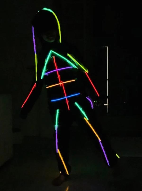 A person wearing a black outfit decorated with brightly colored neon glow sticks in the shape of lines and angles, creating a stick-figure effect against a dark background.