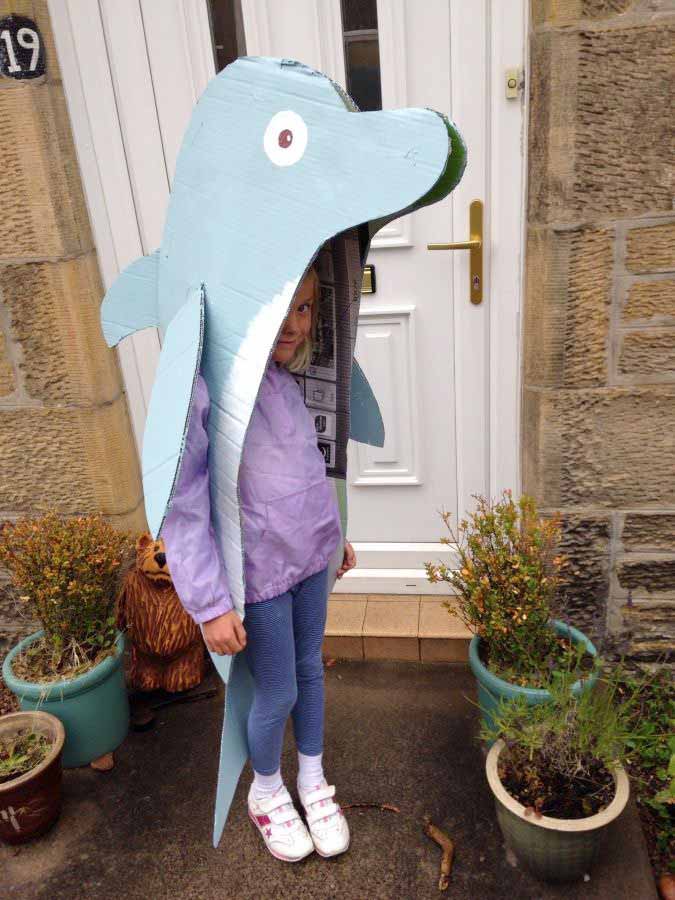 A child stands outside a house wearing a large homemade dolphin costume made from painted cardboard—an adorable pick for under the sea costume ideas. The child's face peeks through the mouth of the dolphin, with potted plants nearby.