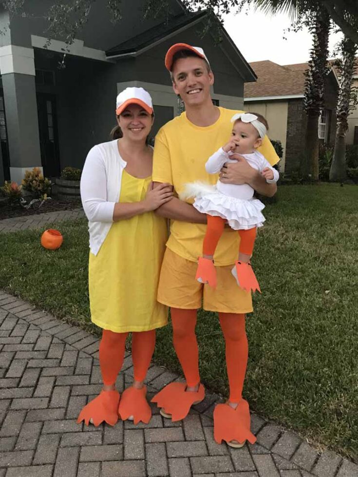 A family of three dressed in matching yellow duck costumes with orange bills, feet, and hats stands outside on a lawn in front of a house. The baby wears a white tutu and a duck bill headband—perfect for fun family costume ideas.