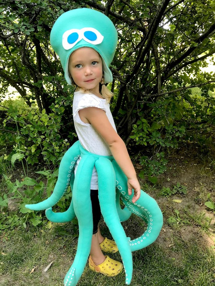 A young child stands outdoors wearing a playful octopus costume with large blue headpiece and long stuffed tentacles, surrounded by green trees and grass.