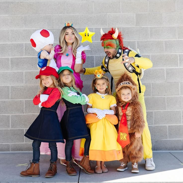 A family dressed in colorful Mario-themed costumes poses against a gray brick wall, including Princess Peach, Bowser, Mario, Luigi, Princess Daisy, Donkey Kong, and Toad.