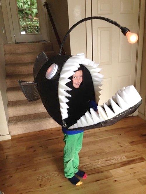A child wearing a creative anglerfish costume with large white teeth, a glowing lure on top, and their face visible inside the fish’s mouth, standing on a wooden floor near a staircase.