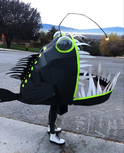 A person wearing a large, homemade anglerfish costume, mostly black with green and white accents, sharp paper teeth, and a glowing lure, stands on a suburban street with trees and hills in the background.