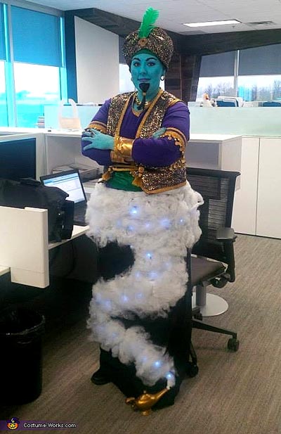 A person dressed as a genie stands in an office, wearing blue face paint, a turban, a gold vest, and a costume resembling a cloud of smoke with lights, giving the appearance of emerging from a lamp.