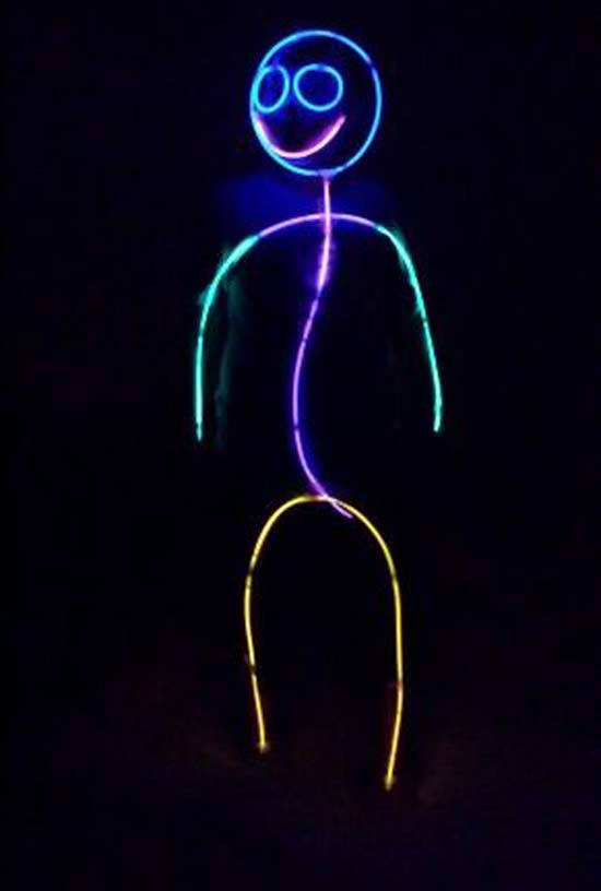 A person is dressed as a glowing stick figure, outlined with colorful neon lights in the dark, forming a smiling face, arms, body, and legs.