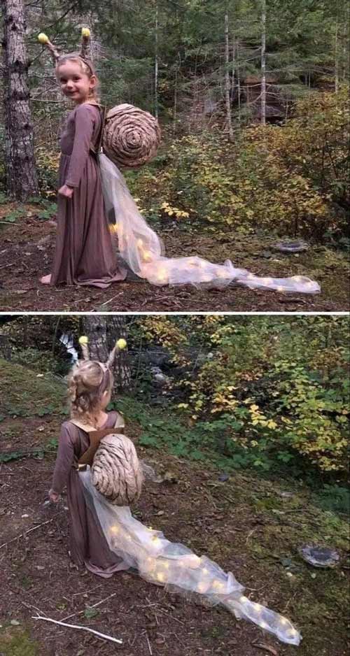 A child in a snail costume stands in a forest. The costume includes a brown dress, a large shell on the back, and a sheer, lit-up train; two antennae with yellow tips are on the child’s head.