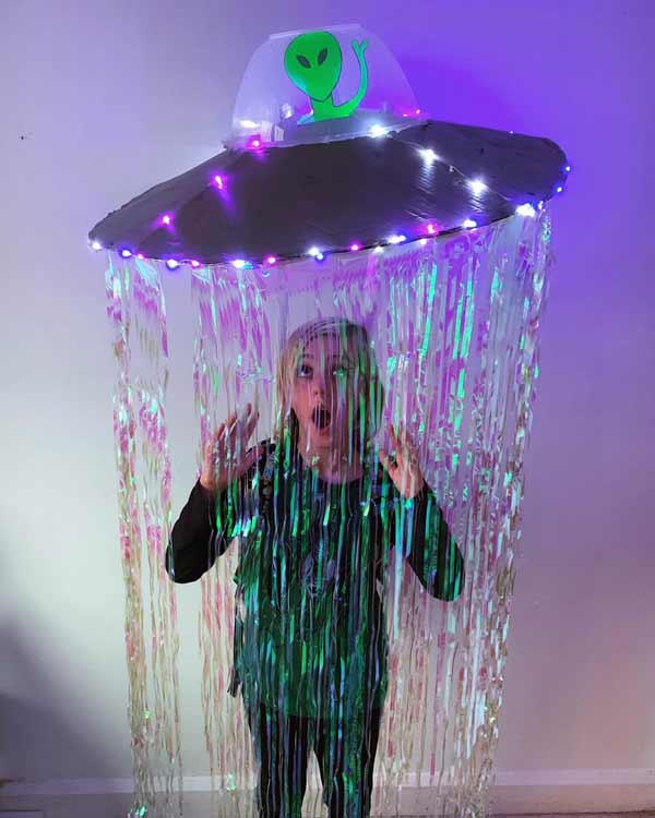 A child stands inside a homemade UFO costume with hanging shimmering streamers, LED lights, and a cardboard sign on top featuring a green alien waving. The child is making a surprised face.