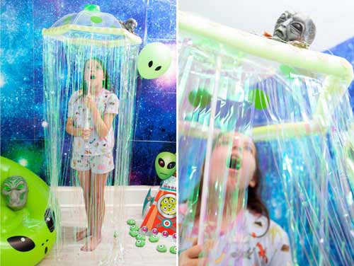 A child stands under a UFO-shaped decoration with hanging streamers, surrounded by alien-themed balloons and toys in a space-themed room. The child looks up at the decoration with awe.