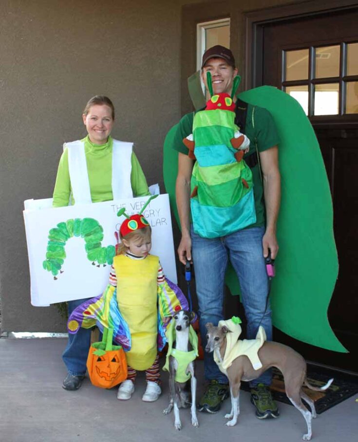 A creative take on family costume ideas: a family and their two dogs dress as characters from "The Very Hungry Caterpillar," with adults as the caterpillar and book, the child as a butterfly, and the pups in adorable matching costumes.