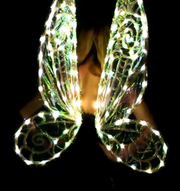 A person with long hair wears large, illuminated fairy wings outlined with glowing lights and swirled patterns, standing against a dark background.