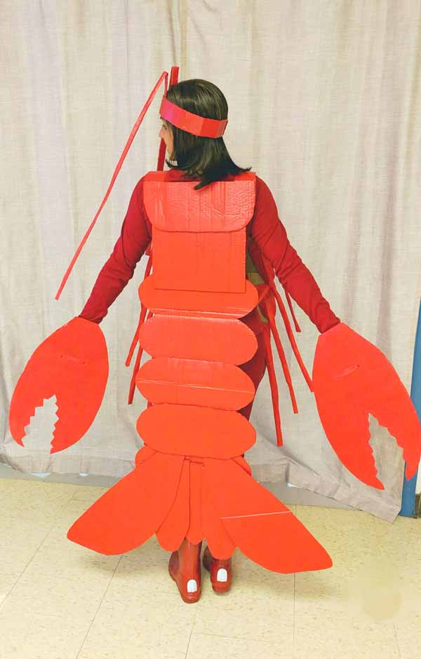 A person wearing a homemade red lobster costume, complete with claws, tail, and antennae, stands facing away from the camera in front of a beige curtain—perfect inspiration for under the sea costume ideas.