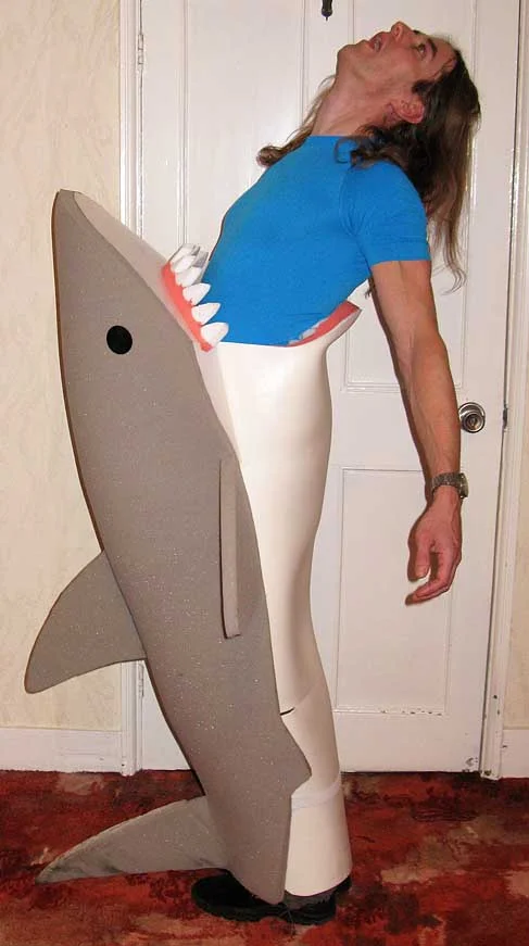 A person in a blue shirt wears a creative costume that makes it seem like a large foam shark is biting them around the waist—perfect for those searching for fun under the sea costume ideas. The person leans back with their mouth open, acting dramatically.