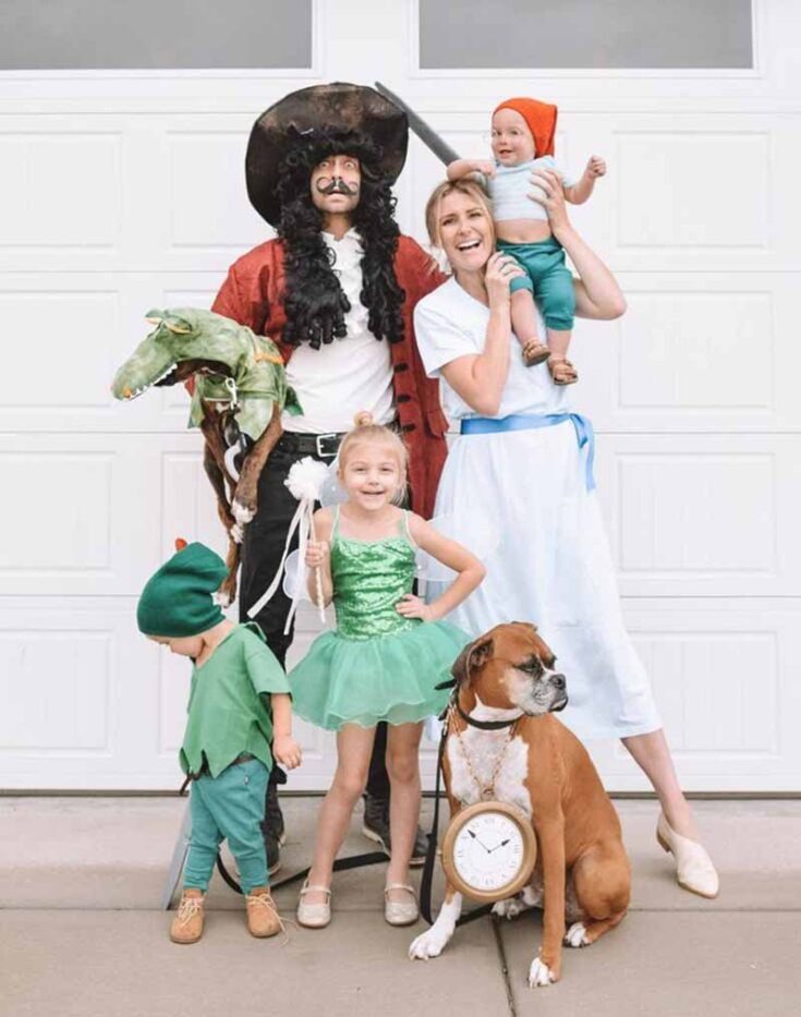 A family dressed in Peter Pan-themed costumes poses in front of a white garage door—one of the best family costume ideas, with adults as Captain Hook and Wendy, kids as Peter Pan, Tinker Bell, and a Lost Boy, and their dog wearing a clock.