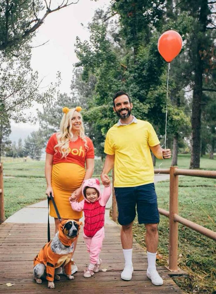 A family in Winnie the Pooh-themed costumes stands on a wooden bridge. Great for family costume ideas, the pregnant woman is Pooh, the man holds a red balloon in a yellow shirt, their child is Piglet, and even their dog joins as Tigger.