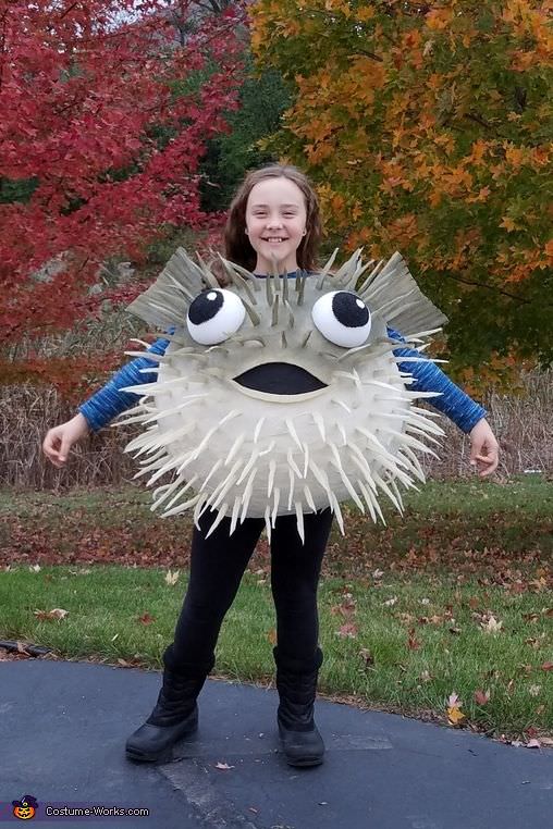 A smiling child stands outdoors in a creative pufferfish outfit—with large eyes and spikes—against autumn trees of red and orange, showcasing fun under the sea costume ideas.
