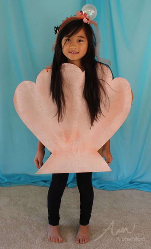 A young girl stands indoors wearing a large seashell-shaped costume and a headband decorated with a bubble and small fish, posing in front of a blue backdrop—perfect inspiration for under the sea costume ideas.