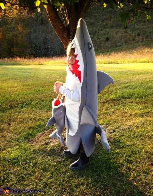 A child stands outdoors in an under the sea costume that makes it look like they are being eaten by a large, gray shark. Holding a trick-or-treat bag, they stand in sunlight on the grassy field behind them.