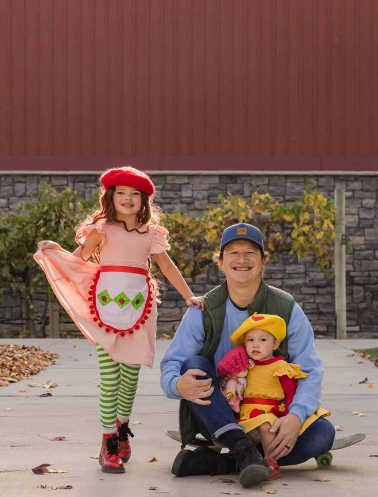 A man sits outside with two young children, all smiling in front of a rustic stone and red barn wall. The older child stands in a strawberry-themed costume while the younger, dressed as a duck, offers adorable family costume ideas.