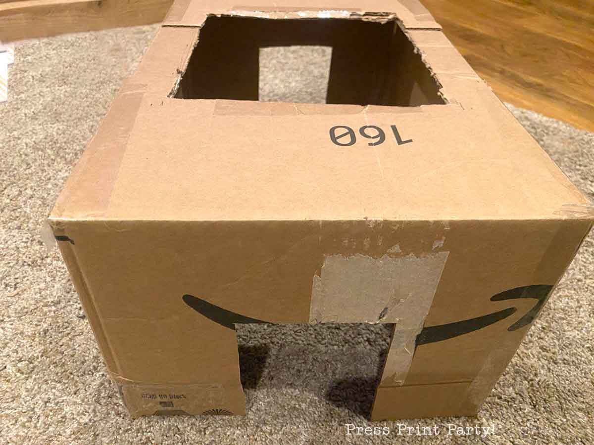 A cardboard box with two rectangular holes cut out, ideal for a tic tac costume, sits on a carpeted floor. The Amazon logo is visible on the front of the box.