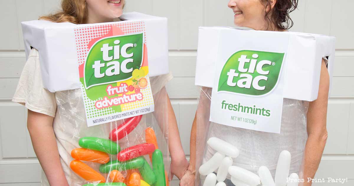 Two people wear creative tic tac costumes—one labeled "fruit adventure" with colorful capsules, the other labeled "freshmints" with white ones. Smiling and holding hands, they bring playful charm to their Tic Tac-inspired outfits.