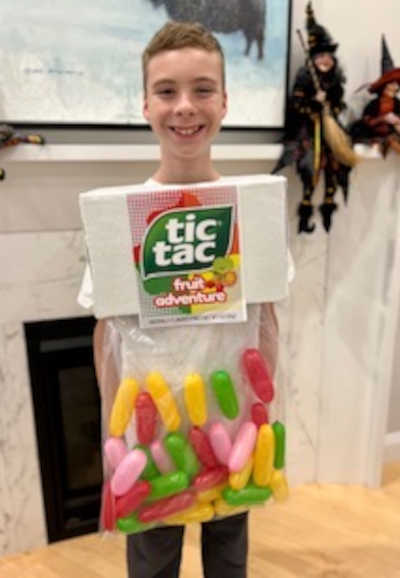 A smiling boy wears a creative tic tac costume, made from a box and plastic bag filled with colorful capsules, standing indoors by a fireplace decorated for Halloween.