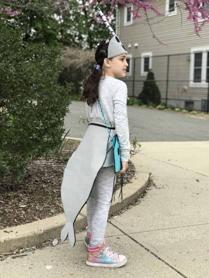 A young girl stands outdoors in a gray narwhal outfit with a horn headband and tail—one of the cutest under the sea costume ideas. She carries a teal purse and wears colorful sneakers near a sidewalk, framed by trees and a nearby building.