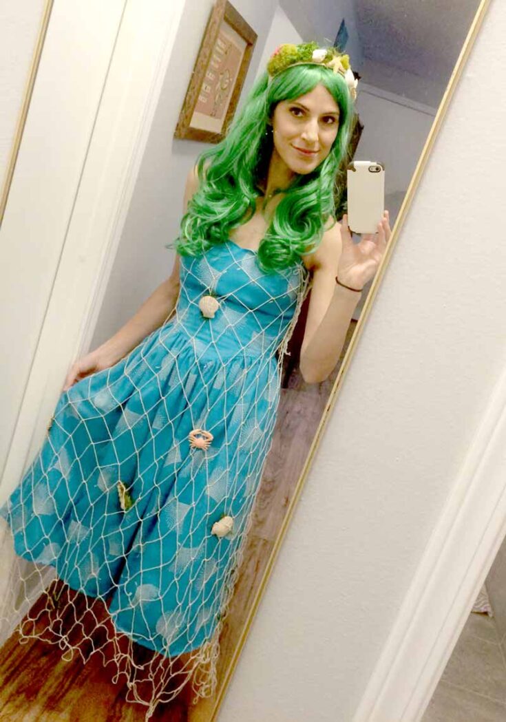 A woman with long green hair wears a blue dress draped with fishnet and seashells, taking a mirror selfie and smiling—one of the cutest under the sea costume ideas.