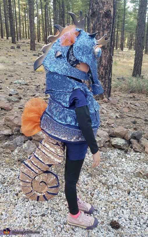 A child outdoors in a sparkly blue and orange seahorse outfit—complete with curled tail, textured body, and googly-eyed headpiece—shows off creative under the sea costume ideas while standing on gravel near trees.