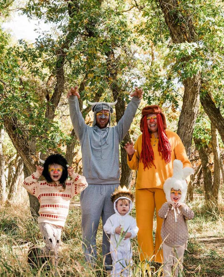 A group of five people, including children and adults, showcase fun family costume ideas as they stand outdoors in a sunlit forest, dressed in whimsical animal and monster costumes, smiling and playfully posing with raised hands.