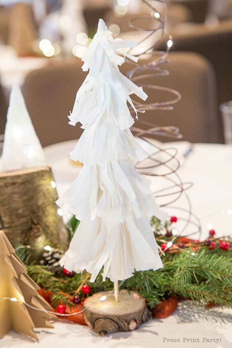 A white, fringed paper Christmas tree centerpiece stands on a wooden base, surrounded by festive greenery, red berries, and other holiday decorations—perfect christmas tree table decor for your holiday gathering. Christmas party centerpiece
