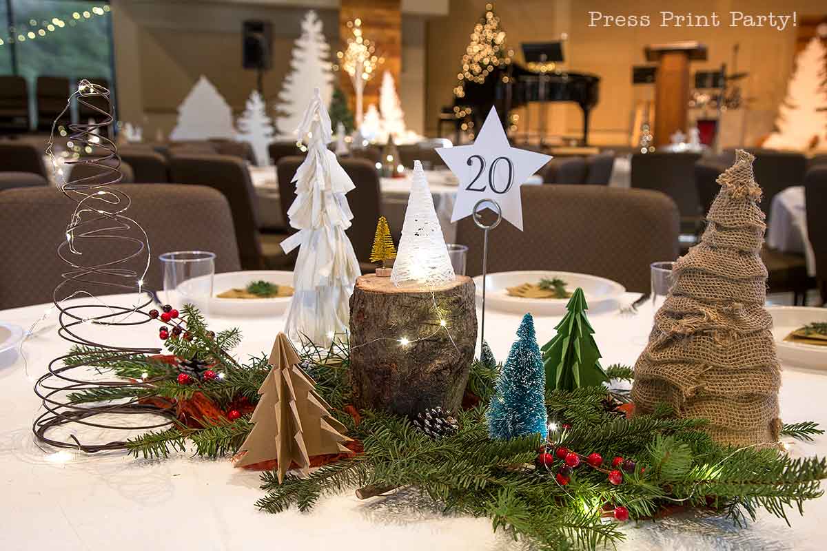 A round table featuring charming christmas tree table decor—miniature trees crafted from paper, wire, burlap, and wood—festive greenery, and red berries encircle a "20" table number; holiday decorations fill the background.