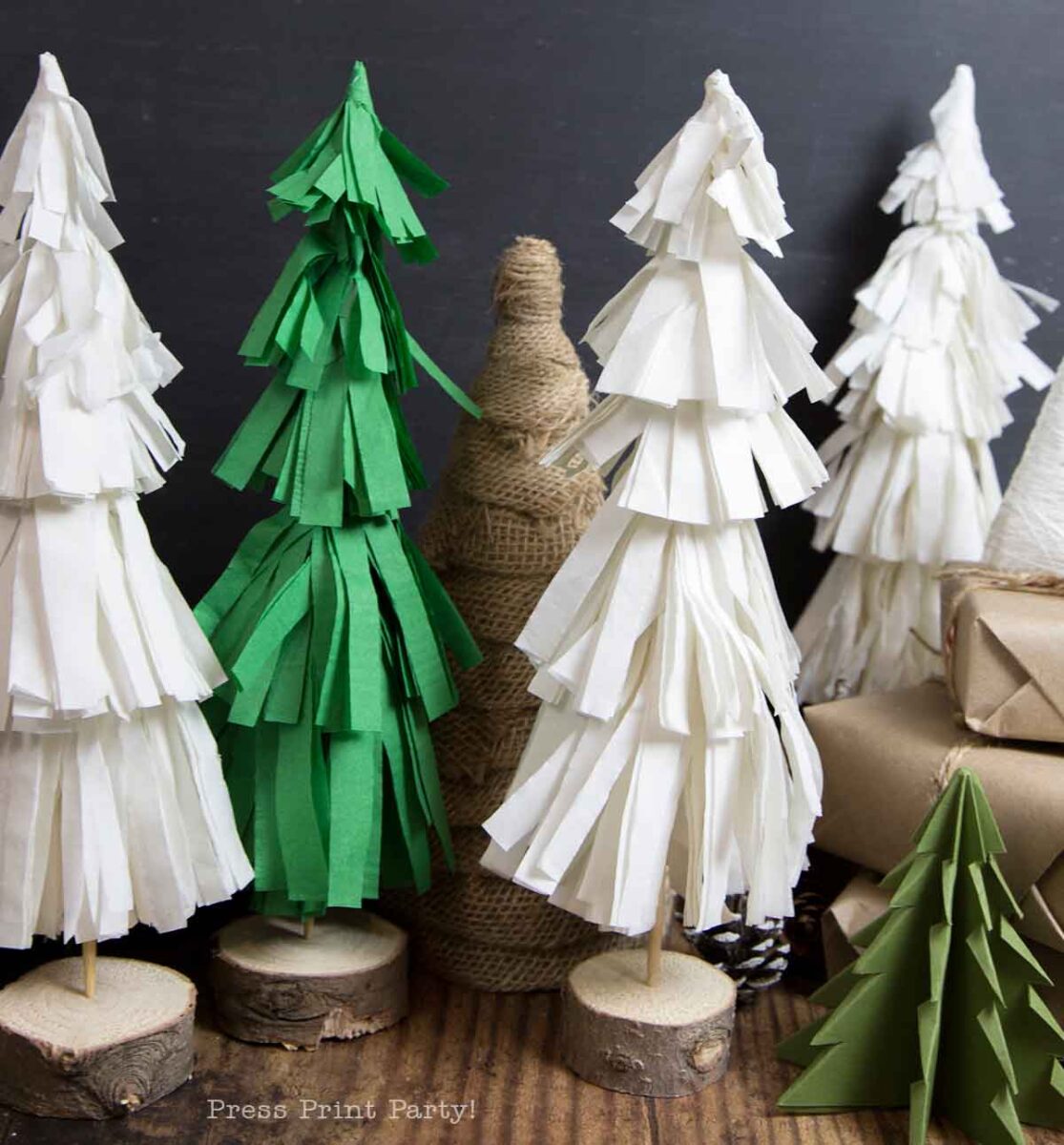 A group of handmade Christmas tree table decor crafted from green and white paper and twine stand on wooden bases, surrounded by wrapped gifts and decorations on a rustic wooden surface.