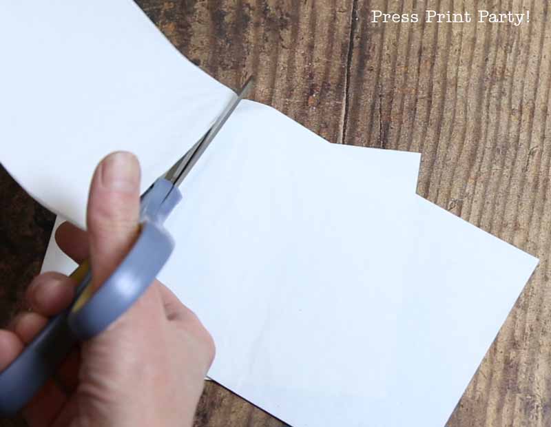 A hand holding scissors cuts a piece of white paper on a wooden surface, perfect for creating Christmas tree table decor. The words "Press Print Party!" appear in the upper right corner.
