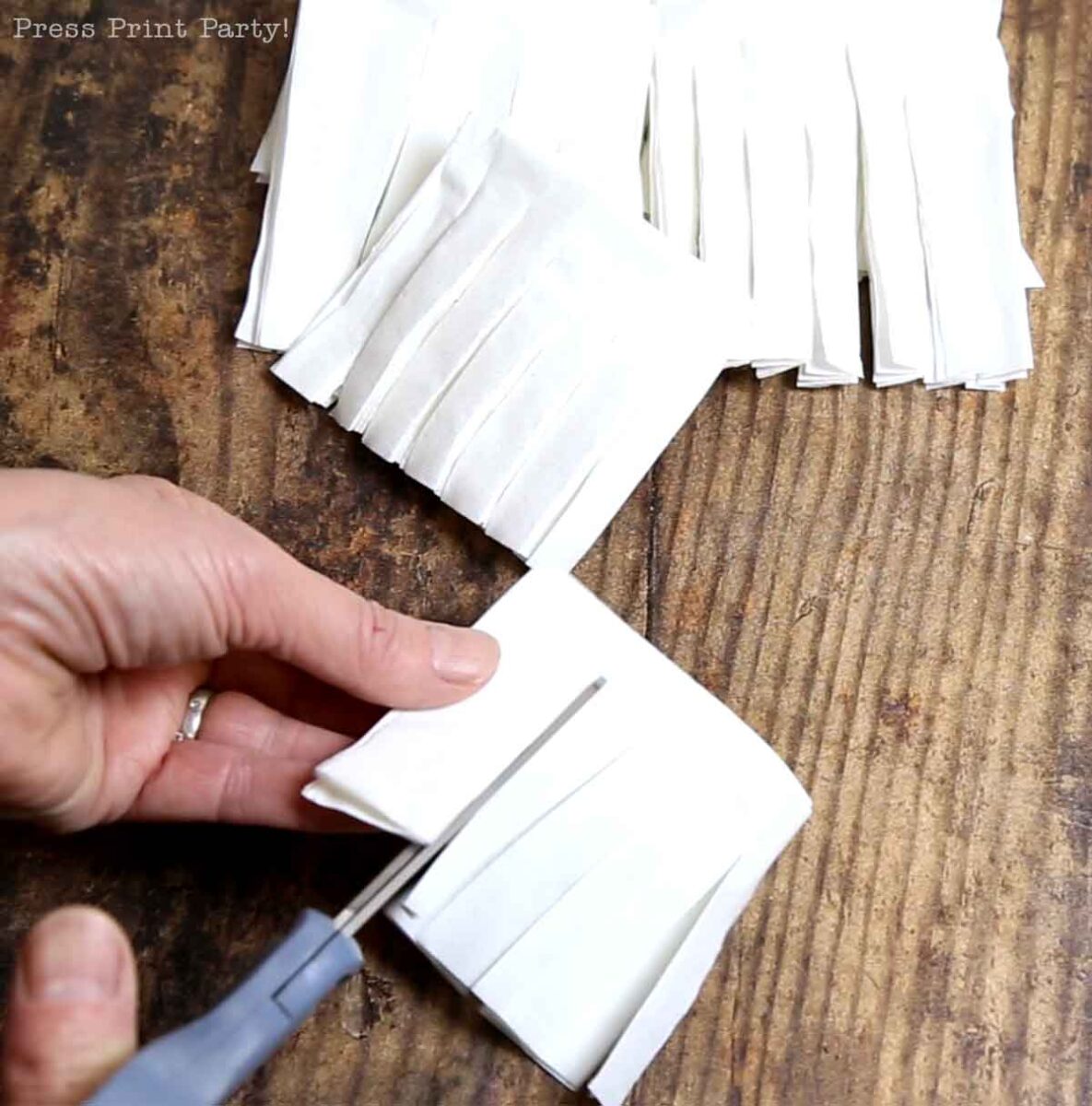 A hand uses scissors to cut fringes into folded white paper on a wooden surface, crafting pieces that can be used for Christmas tree table decor. More folded, fringed papers are stacked in the background, ready for festive arrangements.