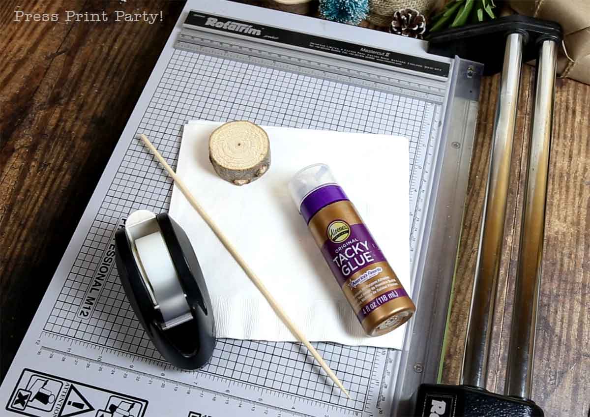 A paper trimmer, clear tape dispenser, tacky glue, wooden stick, wood slice, and a napkin are arranged on a wooden table for a festive christmas tree table decor craft project.