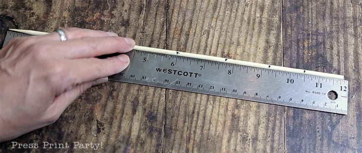 A hand holds a metal ruler marked “Westcott” alongside a wooden dowel on a rustic wooden surface, measuring the dowel—ideal for crafting Christmas tree table decor.