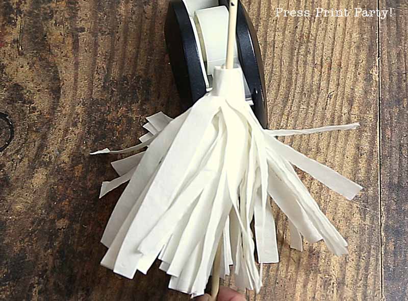 A hand holds a bundle of white paper strips next to a tape dispenser on a wooden surface, crafting a paper tassel perfect for christmas tree table decor.