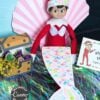 An elf doll sits by a pink paper seashell, wearing a colorful mermaid tail. Beside it is a treasure chest with beads, candy, and an Elf on the Shelf Movie Night Printable Kit card featuring an elf illustration.