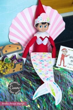 An elf doll sits by a pink paper seashell, wearing a colorful mermaid tail. Beside it is a treasure chest with beads, candy, and an Elf on the Shelf Movie Night Printable Kit card featuring an elf illustration.