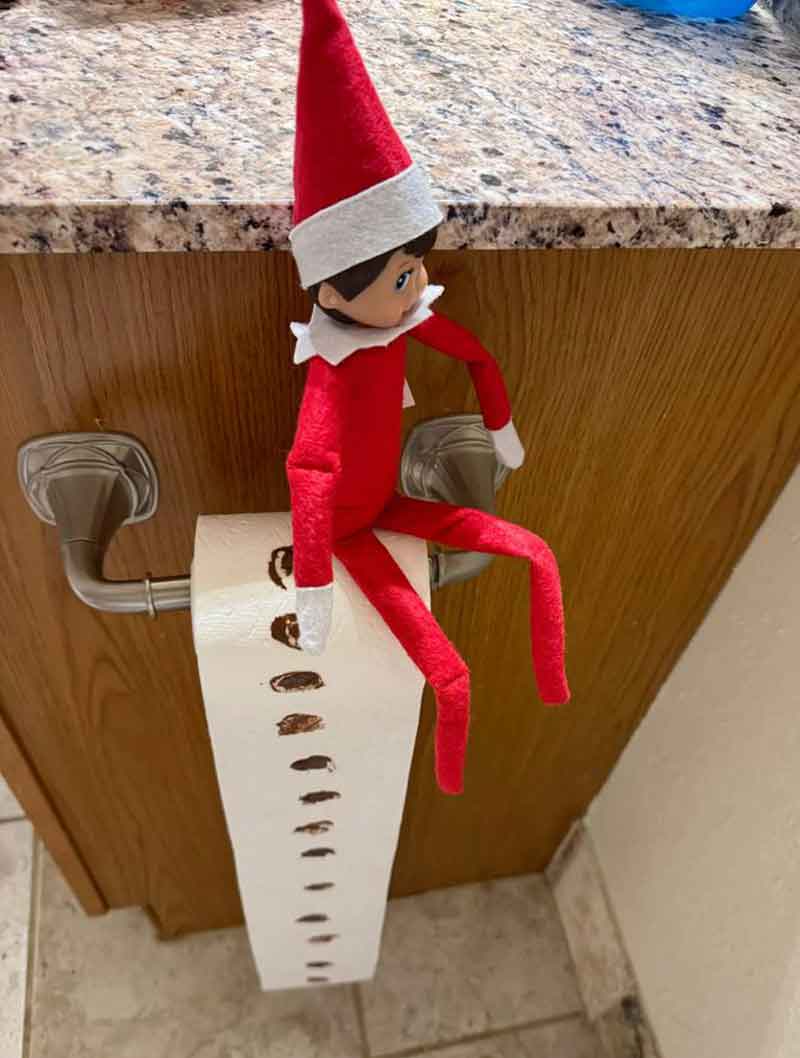 A toy elf in a red outfit sits on a toilet paper roll holder, with the roll unraveled to the floor and brown footprints painted along the toilet paper—perfect for mischievous elf on the shelf poop ideas!.