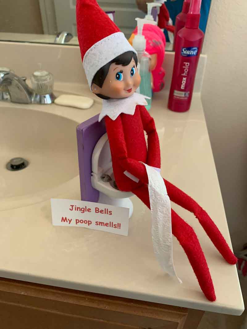An Elf on the Shelf doll sits on a toy toilet in a bathroom, holding toilet paper. A sign reads, "Jingle Bells My poop smells!!" Perfect for those searching for funny elf on the shelf poop ideas, with toiletries scattered on the counter nearby.