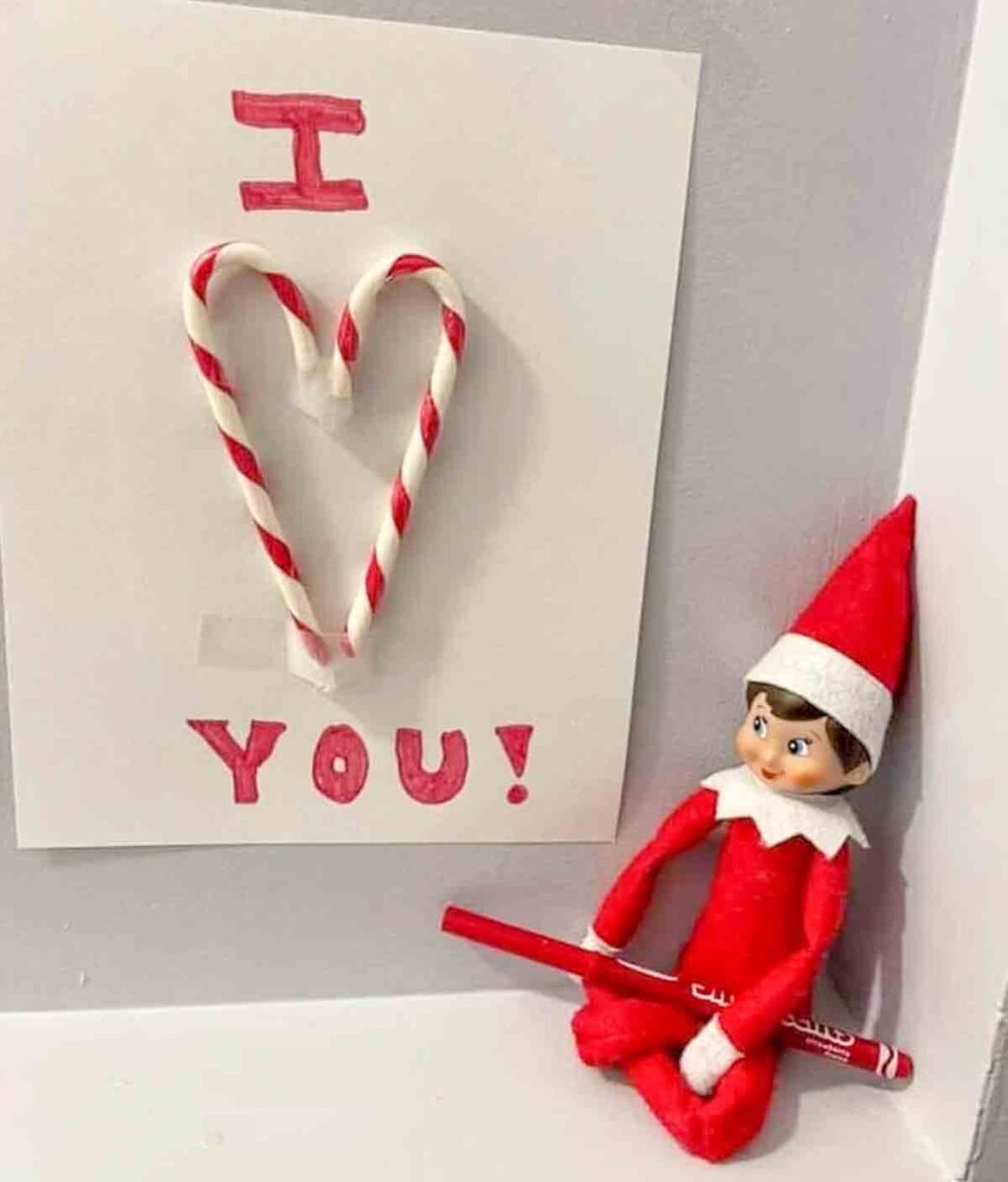 A note with "I โค๏ธ YOU!" uses two candy canes to form a heart shape. Next to it sits an Elf on the Shelf doll holding a red marker.