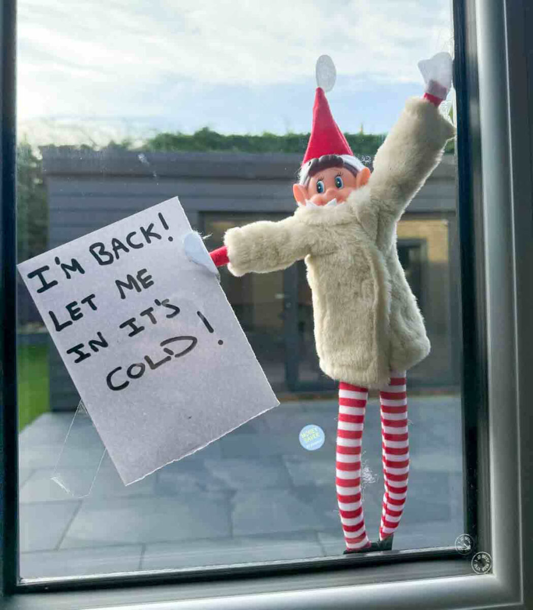A toy elf in a fur coat and striped leggings is stuck to a window, holding a handwritten sign that says, "I'M BACK! LET ME IN IT'S COLD!" A building and trees are visible outside.
