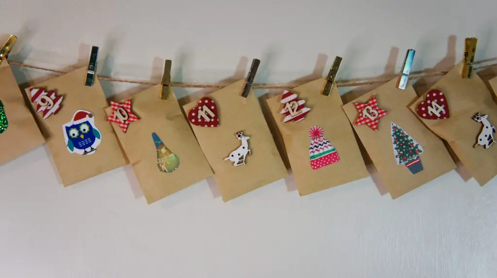 Small brown envelopes decorated with holiday-themed stickers and numbers are clipped to a string with clothespins, creating a festive homemade advent calendar hanging against a white wall.